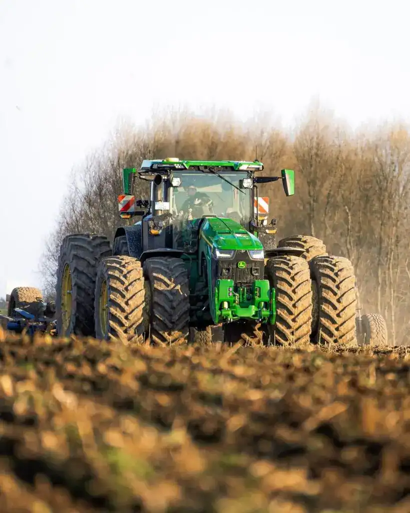 Green John Deere tractor working on farmland in spring, large tires, modern agricultural machinery, driving through tilled soil, environmentally friendly farming equipment, crop cultivation, precision agriculture.