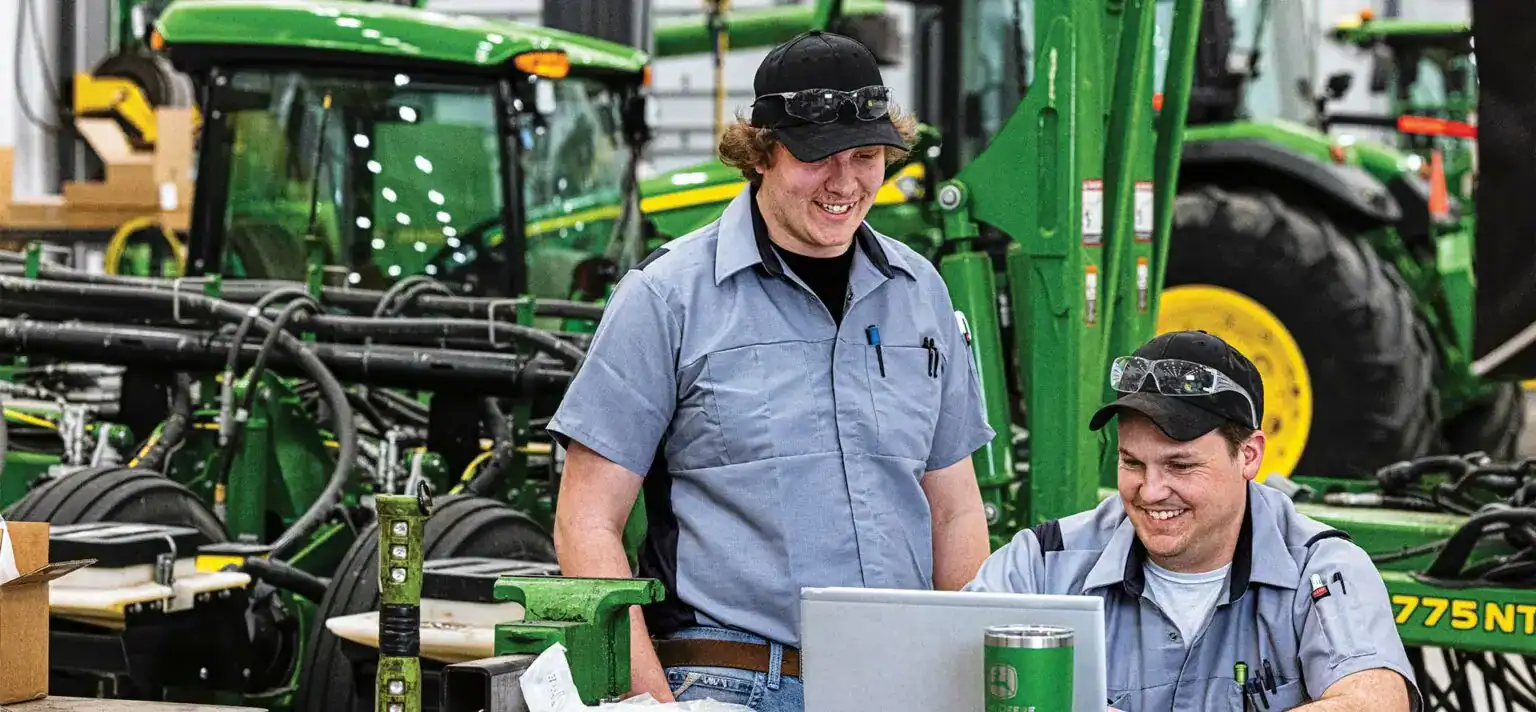 High-quality green John Deere agricultural machinery at JCE AG, with two smiling technicians working together in a well-equipped farm equipment dealership.